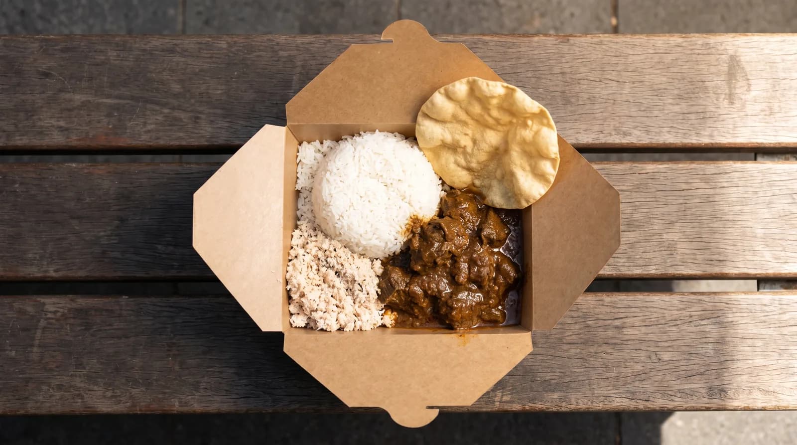 Overhead view of a kraft takeaway box with rice, curry, sambol and a papadum.